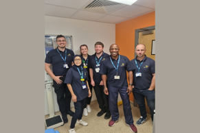 Group of six staff members wearing navy blue polo shirts and ID badges standing together in a clinical setting. The group is photographed in front of an orange wall and doorway with clinical equipment visible in the room.