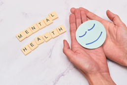 Hands holding a paper circle with a simple smiling face, next to Scrabble-style tiles spelling Mental Health.