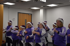 A group of people wearing purple and white festive outfits and hats are standing indoors, performing coordinated hand movements as part of the Learning Disabilities Research event.