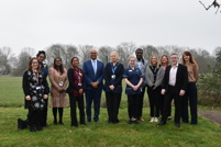 A group of staff stand in a line on grass outdoors, with trees and open fields in the background, posing for a group photo.