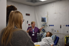 A dummy lies on a bed with a staff member in a maroon uniform speaking and observers looking on