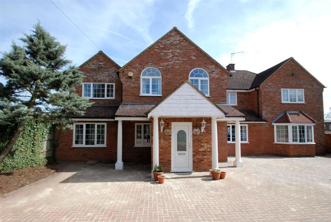 A large red‑brick detached house with white window frames, a central porch supported by columns, and a paved driveway in front. A small tree stands to the left.
